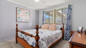 A bedroom with a wooden bed, a ceiling fan, a window with curtains, and a framed picture on the wall. - Blue Meadow - Pet Friendly (Evans Head)