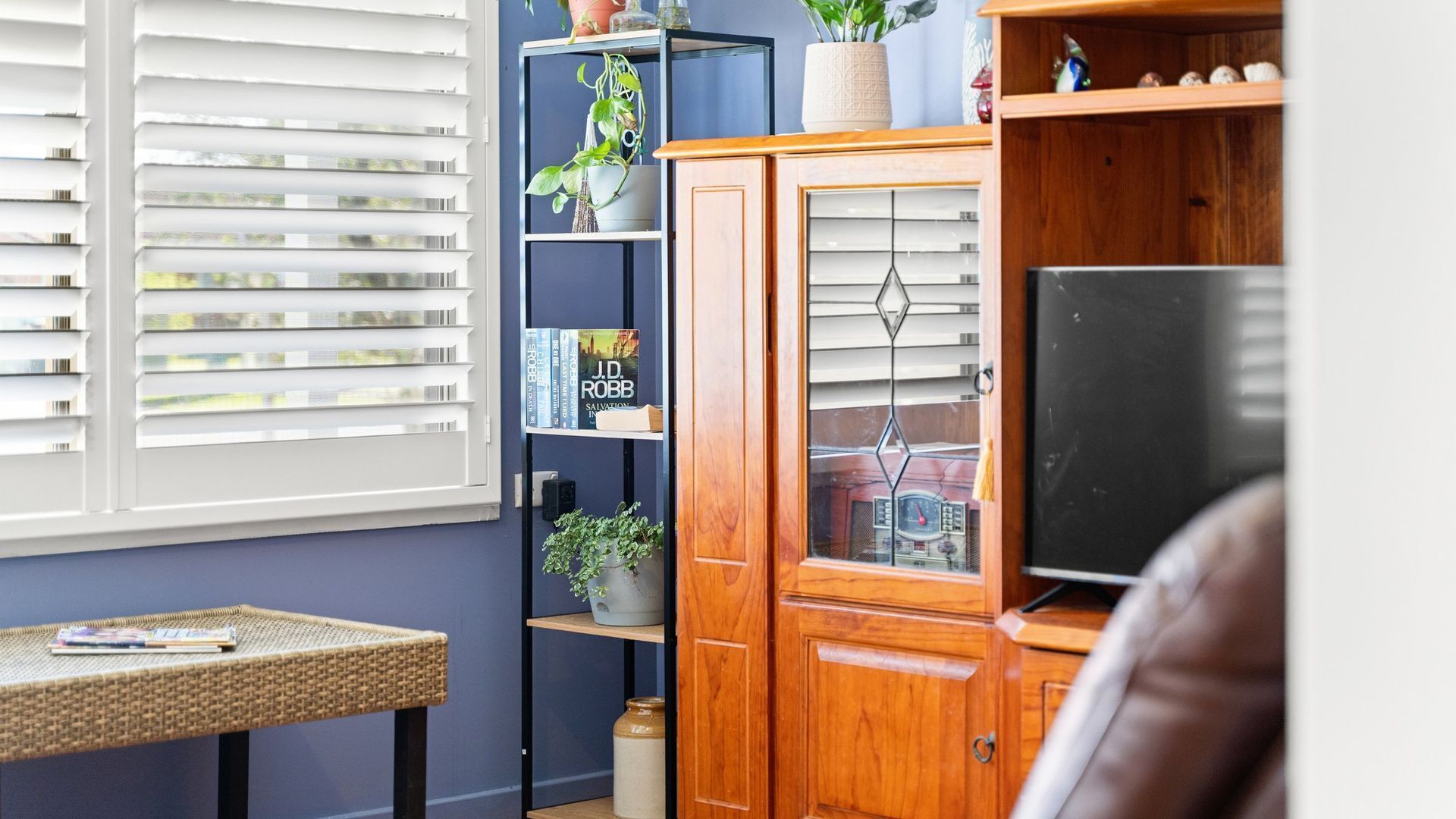 A room with a window with blinds, a bookshelf, a wooden cabinet with a television, and a wicker table.