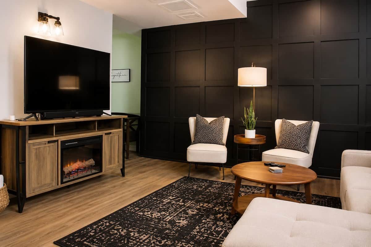 A modern living room with a flat-screen TV, two white armchairs, a coffee table, a fireplace, and a dark wall paneling.
