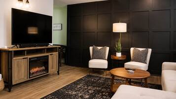 A modern living room with a flat-screen TV, two white armchairs, a coffee table, a fireplace, and a dark wall paneling.