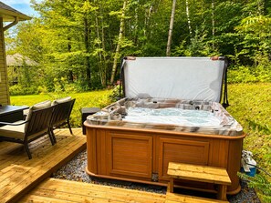 A hot tub on a wooden deck with a bench and table nearby, surrounded by greenery.