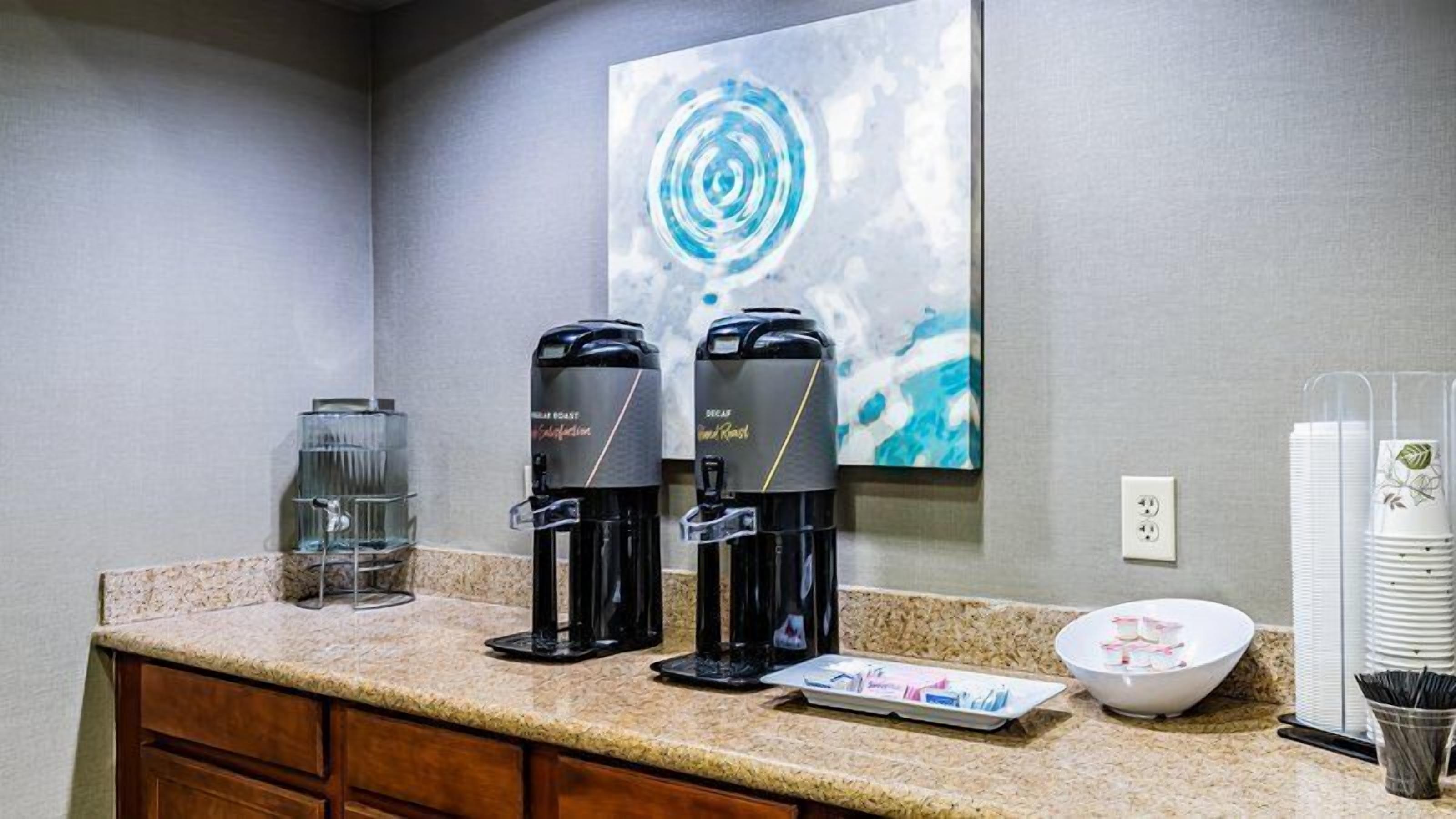 A hotel kitchenette with two coffee makers, a glass water dispenser, a bowl, and stacked plates.