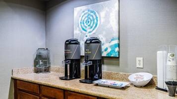 A hotel kitchenette with two coffee makers, a glass water dispenser, a bowl, and stacked plates.
