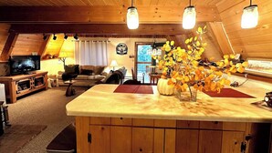 A cozy interior with a wooden ceiling, a central island with a vase of yellow flowers, a television, and a dining area.