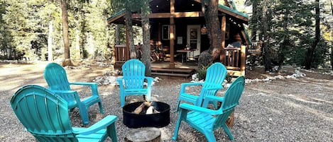 Eine Hütte mit einer Terrasse, umgeben von Bäumen, und vier türkisblauen Adirondack-Stühlen, die um eine Feuerstelle angeordnet sind.