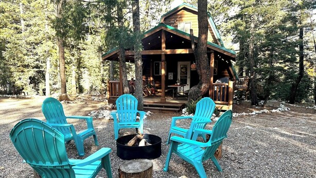 A cabin with a deck, surrounded by trees, and four turquoise Adirondack chairs arranged around a fire pit.