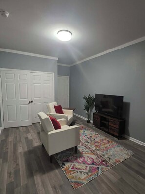 Ein Wohnzimmer mit einem Flachbildfernseher, zwei weißen Sesseln mit roten Kissen, einem farbenfrohen Teppich und Holzboden.