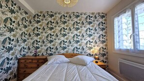 Ein Schlafzimmer mit floraler Tapete, einem hölzernen Bett, einem Nachttisch mit Lampe und einem Fenster mit Vorhängen.