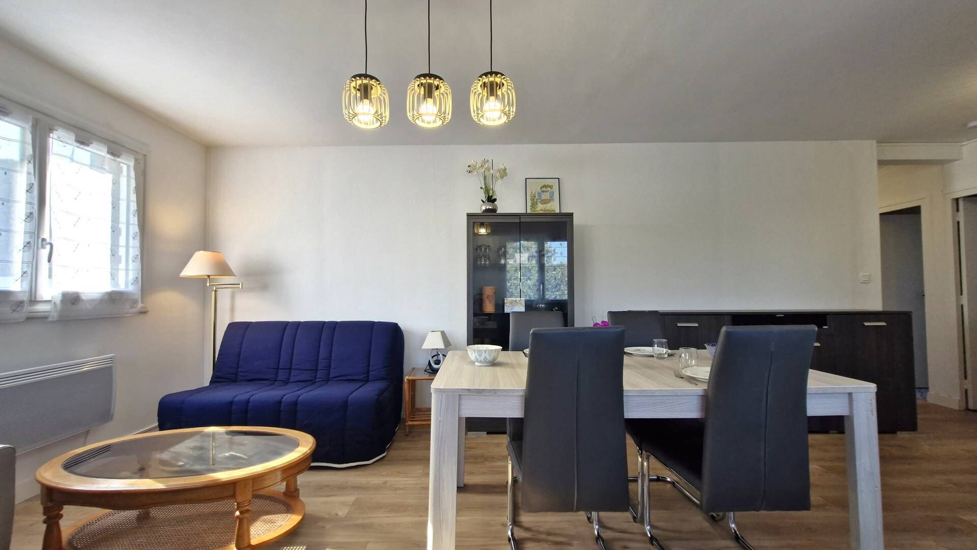 A modern living room with a sofa, a dining table, and a glass coffee table. There are three pendant lights above the table.