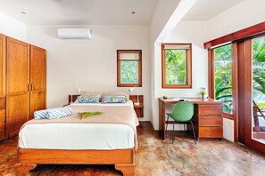 A bedroom with a wooden bed, a desk, and a chair, a window with a view of greenery, and an air conditioning unit.