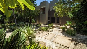 A modern building with a flat roof, surrounded by a garden with various plants and a gravel pathway. - Casa Amorosa: Private Villa with Pool Near Beach & Surf Break playa grande (Playa Grande)