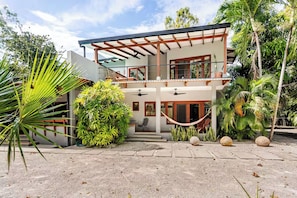 Una casa moderna de dos pisos con un porche cubierto, rodeada de vegetación tropical.