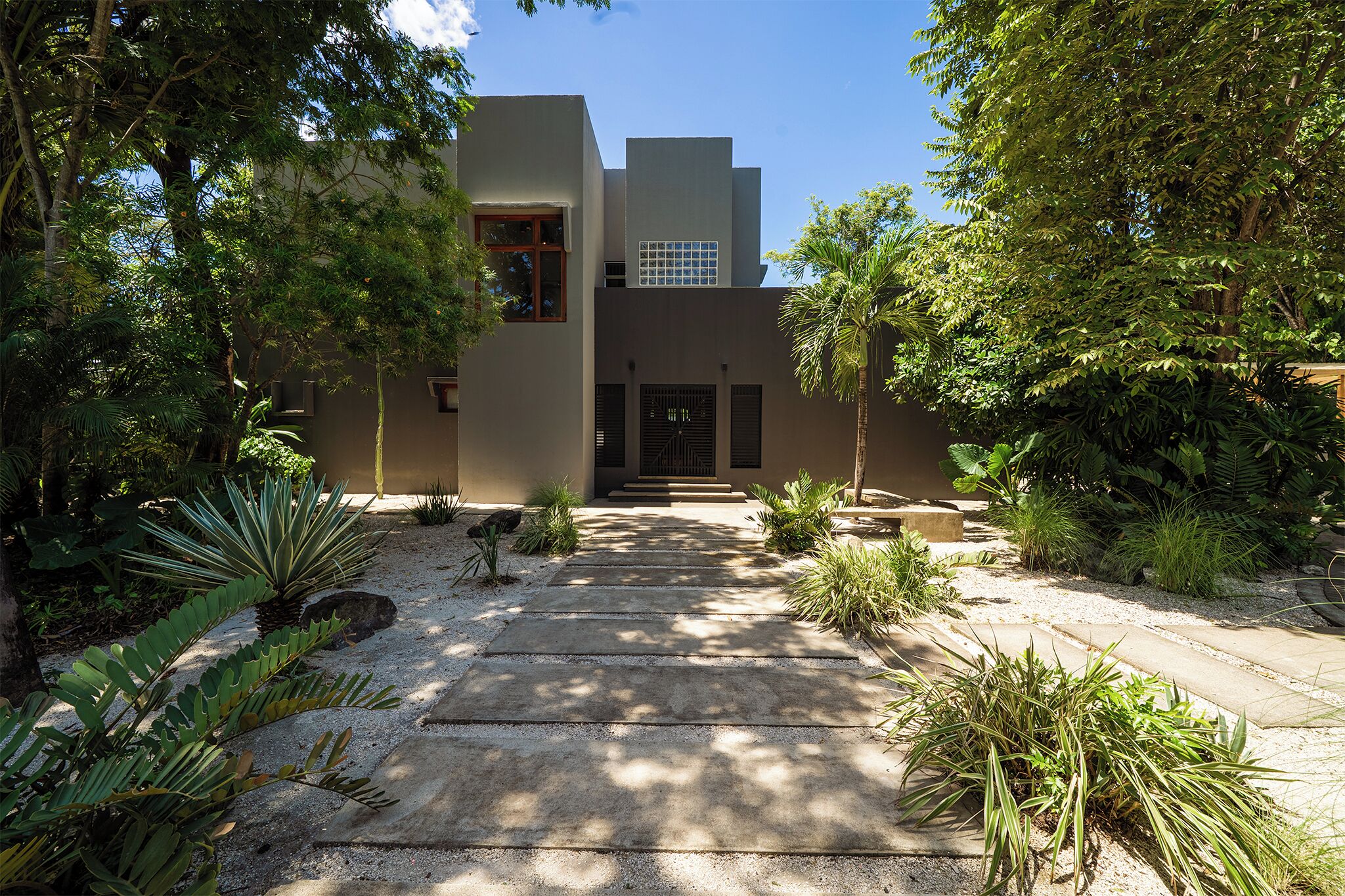 A modern building with a concrete pathway leading to a gated entrance, surrounded by various plants and trees.
