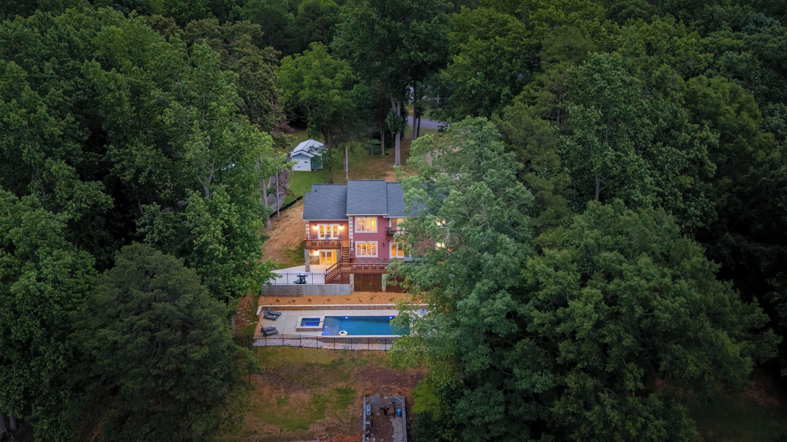 A two-story house surrounded by dense forest, with a visible swimming pool and a fenced yard.