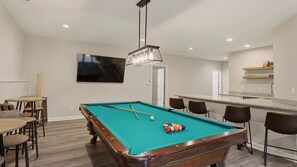 Een pooltafel met stokken en ballen, een bar met krukken, een televisie aan de muur en een deur naar een andere ruimte.