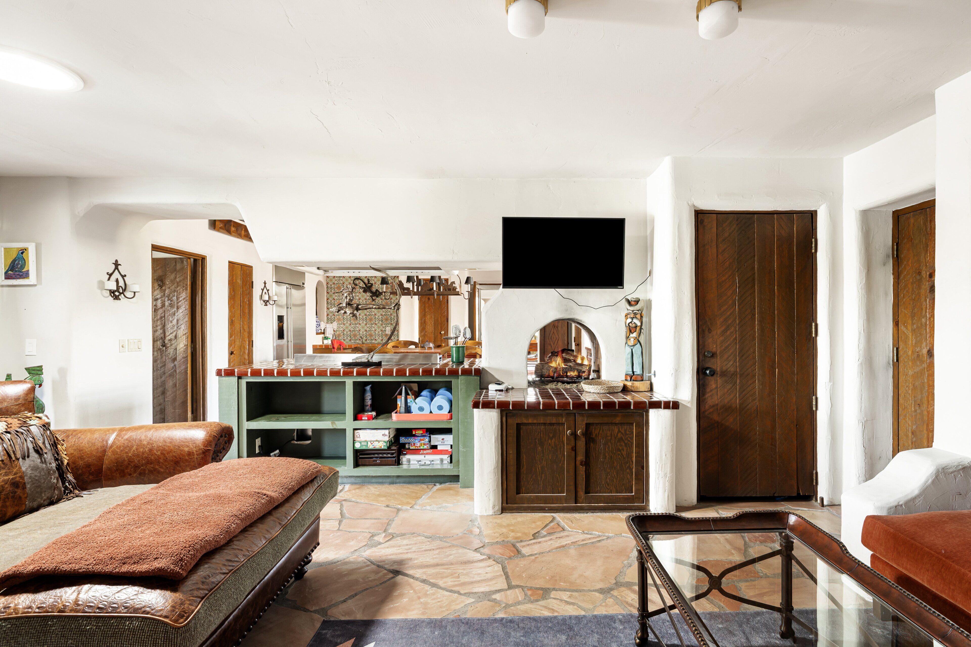 A living room with a sofa, a coffee table, a TV mounted on the wall, and a kitchen area with a counter and cabinets.