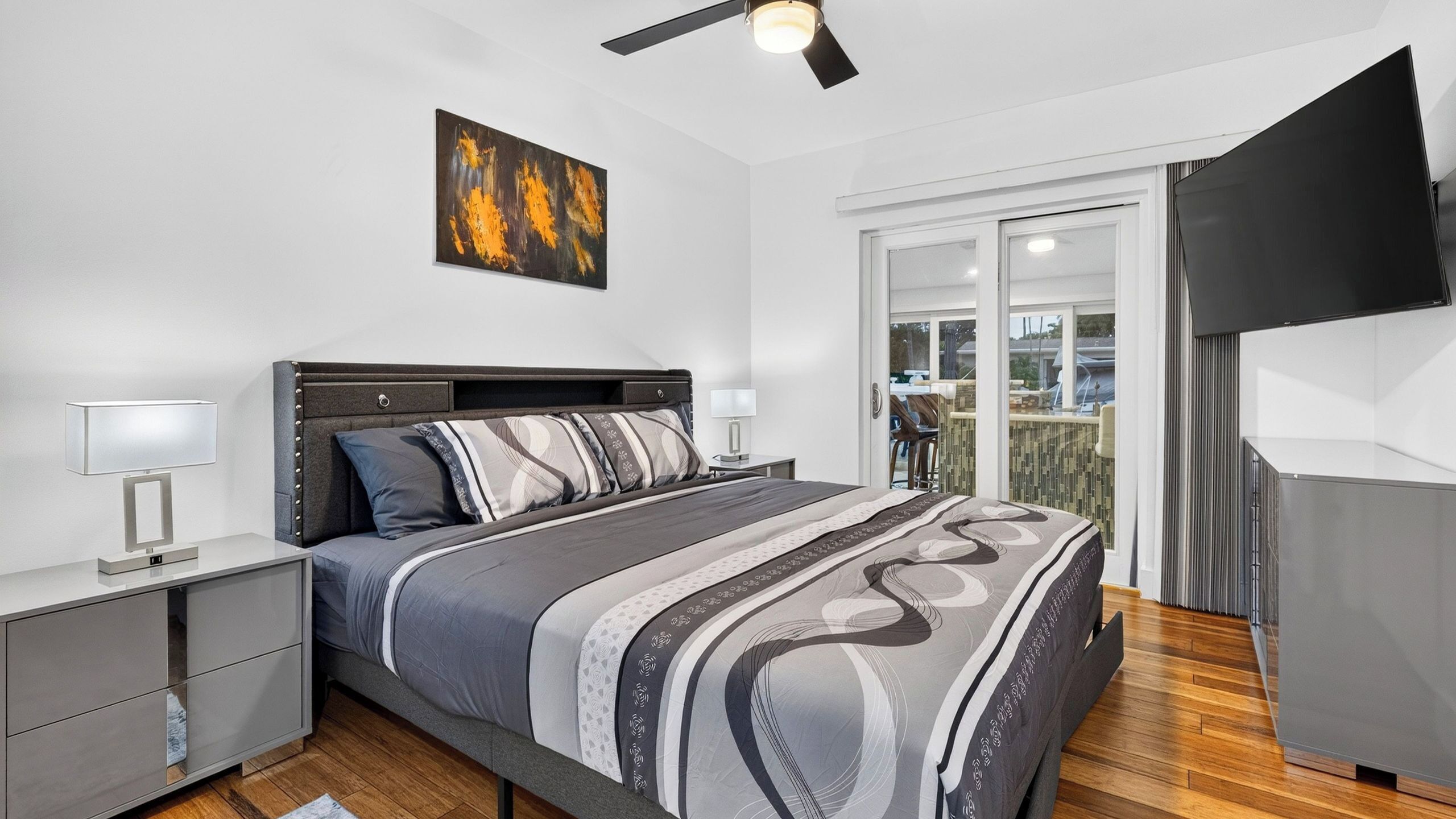 Une chambre moderne avec un lit, des tables de chevet, un ventilateur de plafond, un tableau et une vue à travers une porte vitrée.