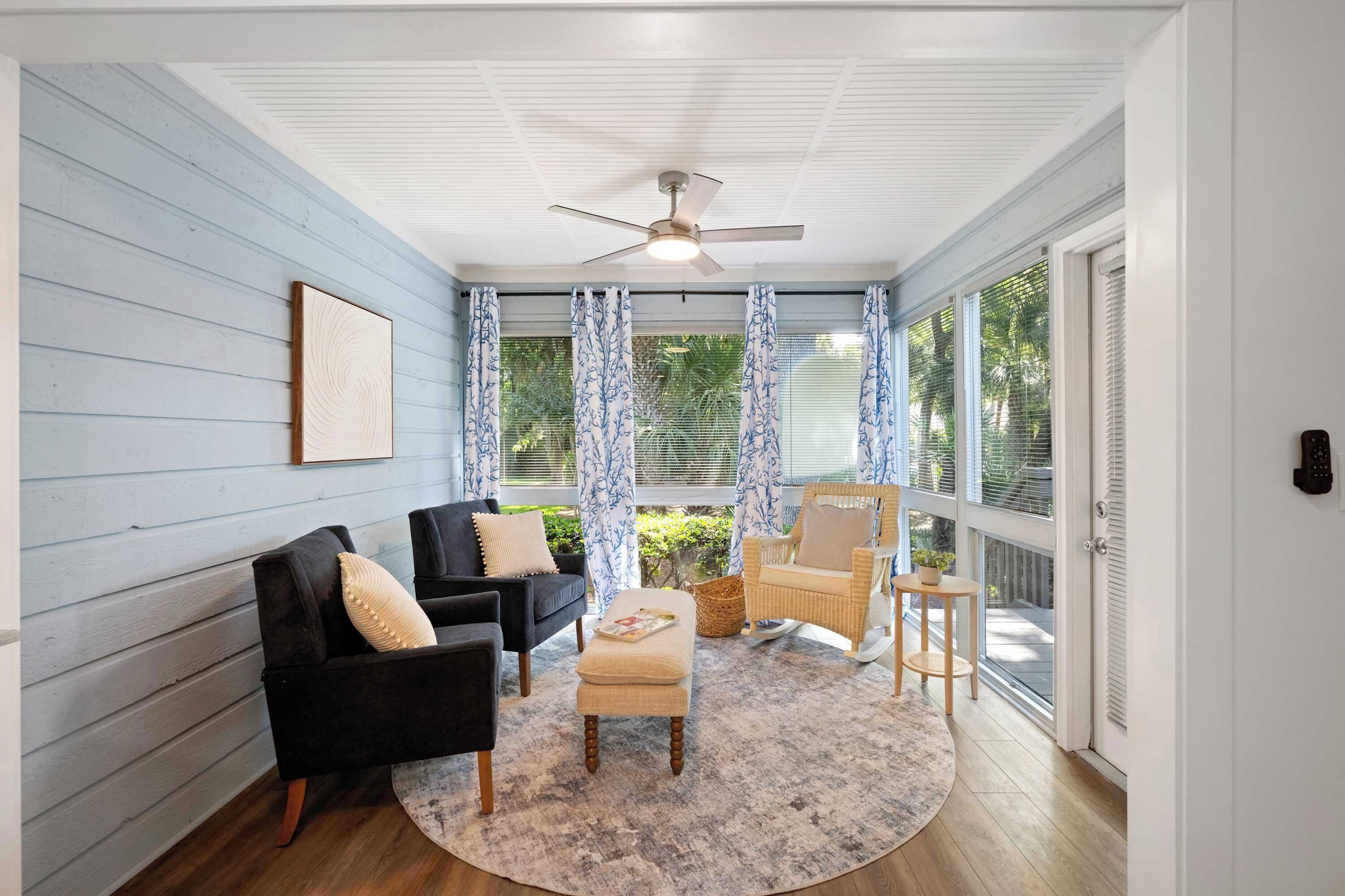 A sunroom with a ceiling fan, a black sofa, a beige armchair, a round rug, and a wicker chair.