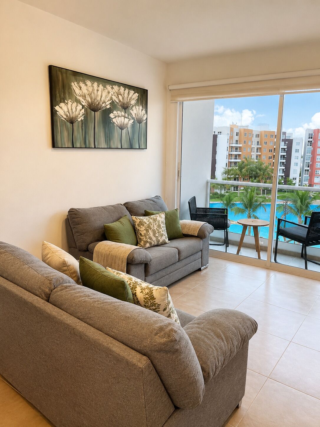 Uma sala de estar com sofá cinza, um quadro de flores e vista para uma piscina e edifícios.
