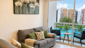 Un soggiorno con un divano grigio, un quadro con fiori e vista su una piscina e degli edifici.