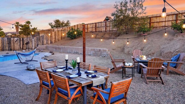 An outdoor patio with a pool, wooden chairs, a table set for dining, string lights, and a wooden fence. - The Light of the Desert by Avantstay Pool + Firepit + BBQ Mins to National Park + DT Yucca Valley (Yucca Valley)