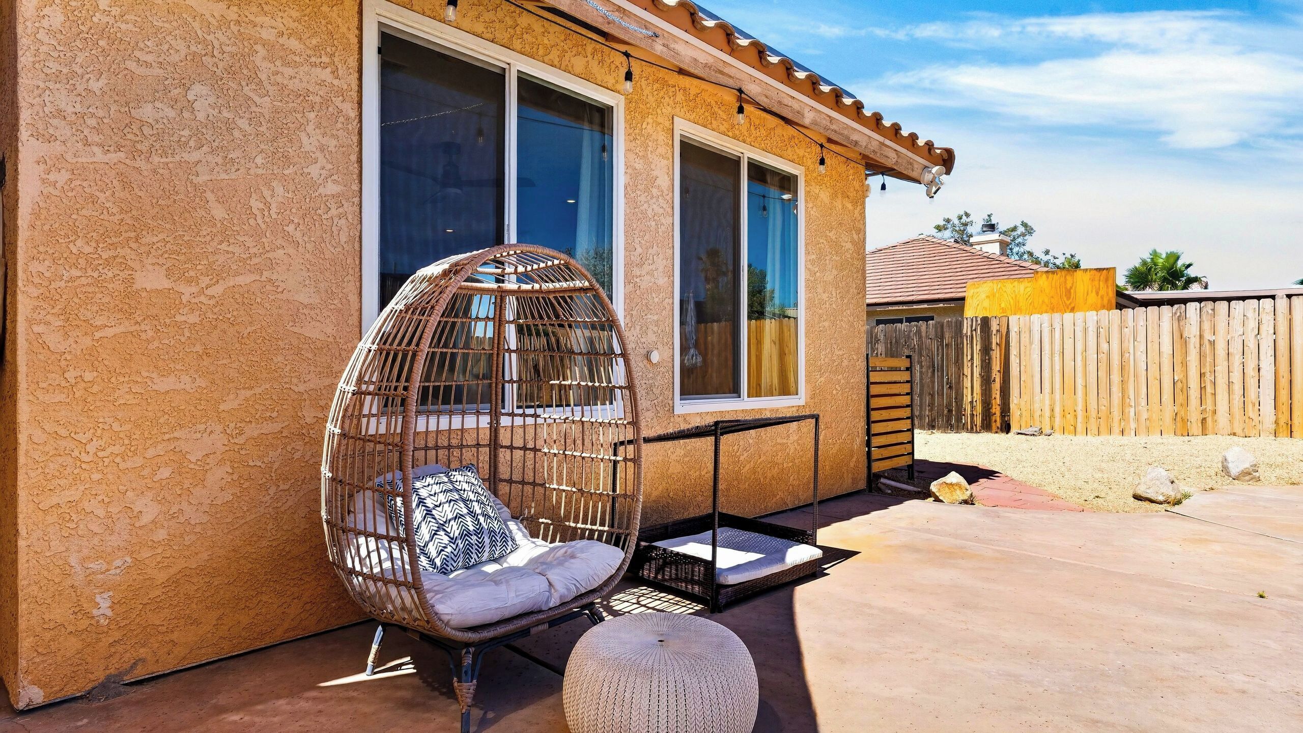 Une maison avec un patio, une chaise en osier avec des coussins et une clôture en bois.