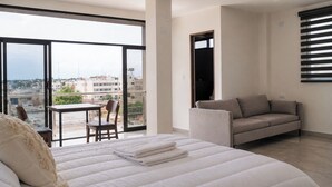 Un dormitorio con una cama grande, un sofá, una mesita y una silla. Tiene un balcón con vistas a edificios.