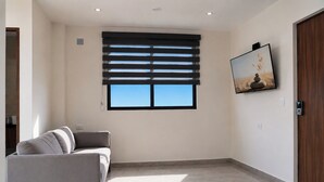 Una habitación de hotel moderna con sofá, cama, ventana con persianas, un cuadro en la pared y una puerta de madera.