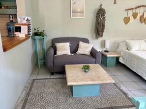 A living room with a grey sofa, a coffee table with a plant, a teal side table, and a white bed with patterned bedding. - Charming studio apartment, centrally located with a garden. (Cozumel)