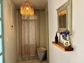 Een badkamer met douchegordijn, een hanglamp, een spiegel met decoratieve lijst, een klein plankje met een fles en een vaas bloemen, en een afvalbak.