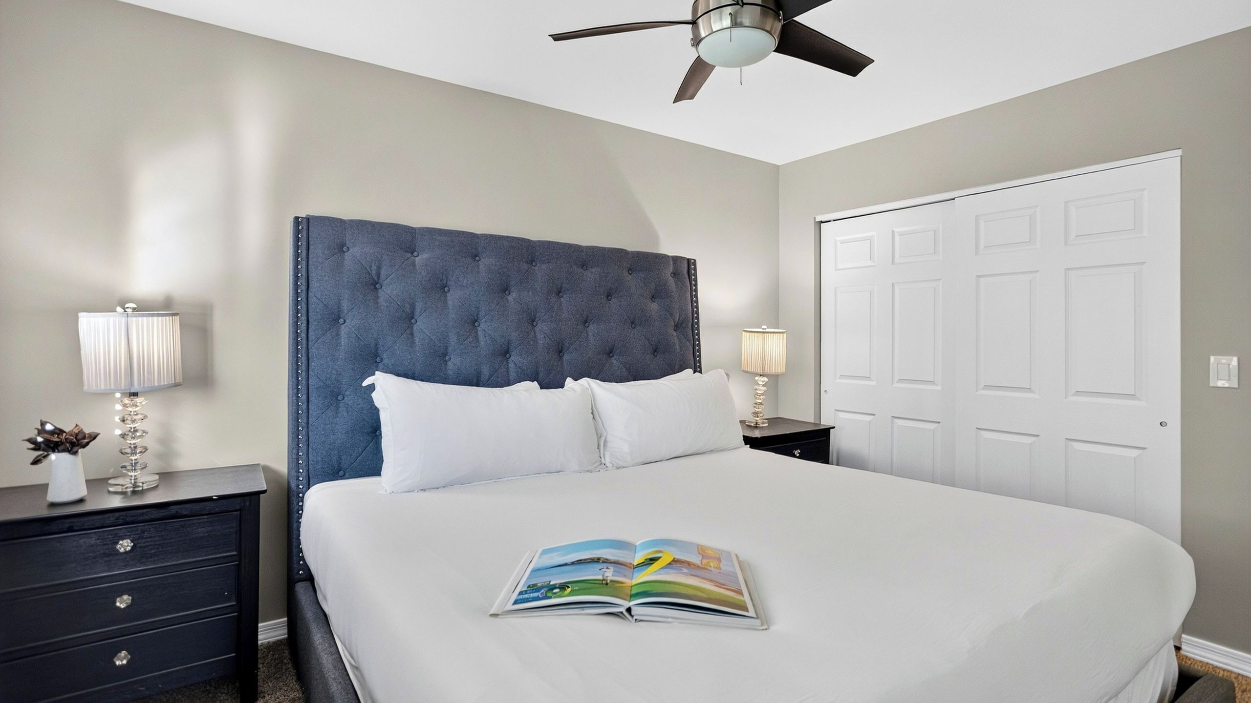 A neatly made bed with a book, a dark headboard, bedside lamps, a nightstand, and a door.