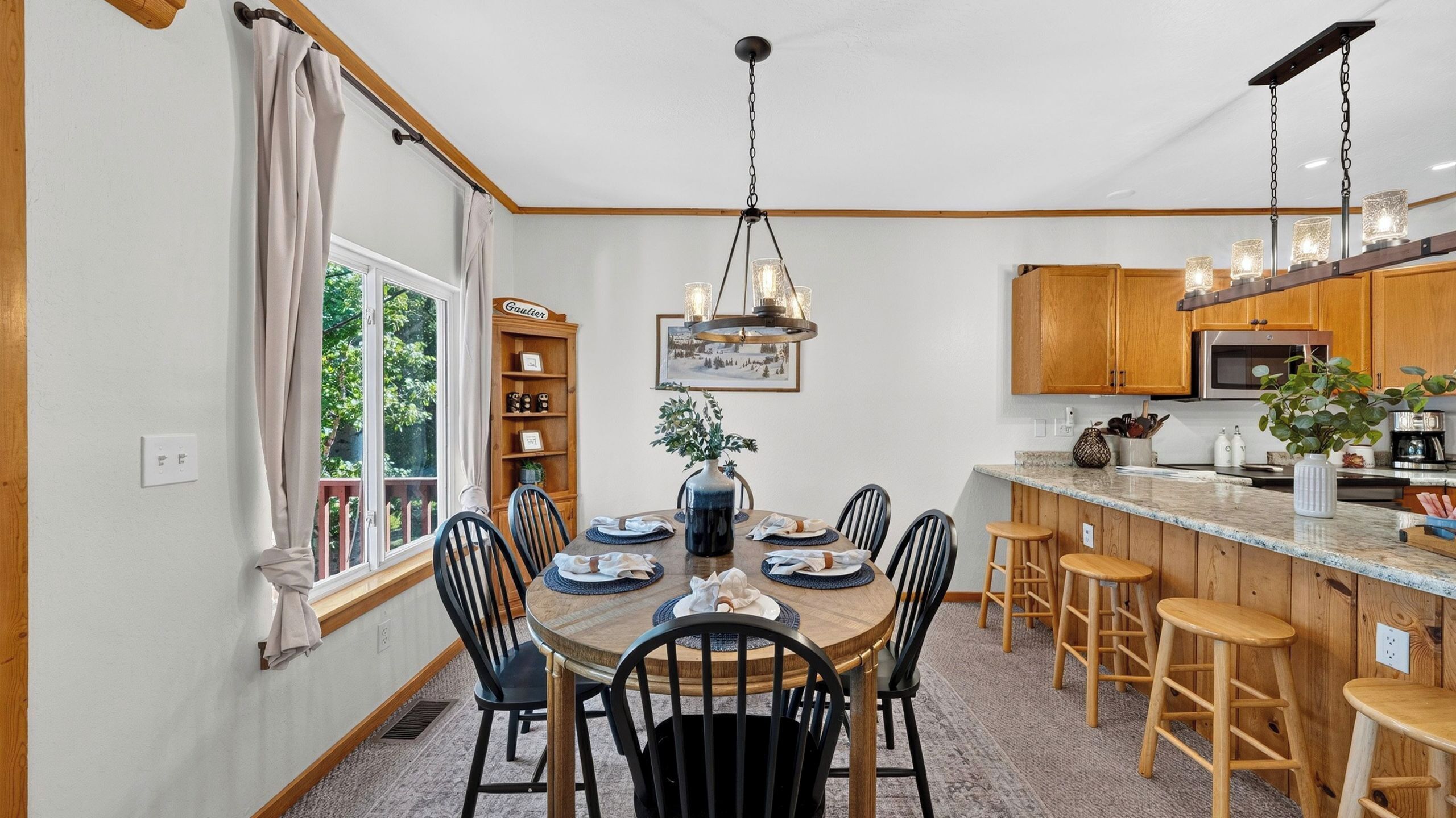 A dining area with a wooden table, chairs, a chandelier, and a kitchen with wooden cabinets and a microwave.