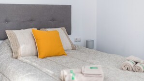 Una cama bien hecha con cabecera gris, un coj ín amarillo brillante y toallas enrolladas a un lado.
