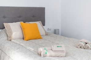 A neatly made bed with a grey headboard, a bright yellow pillow, and rolled towels on the side.
