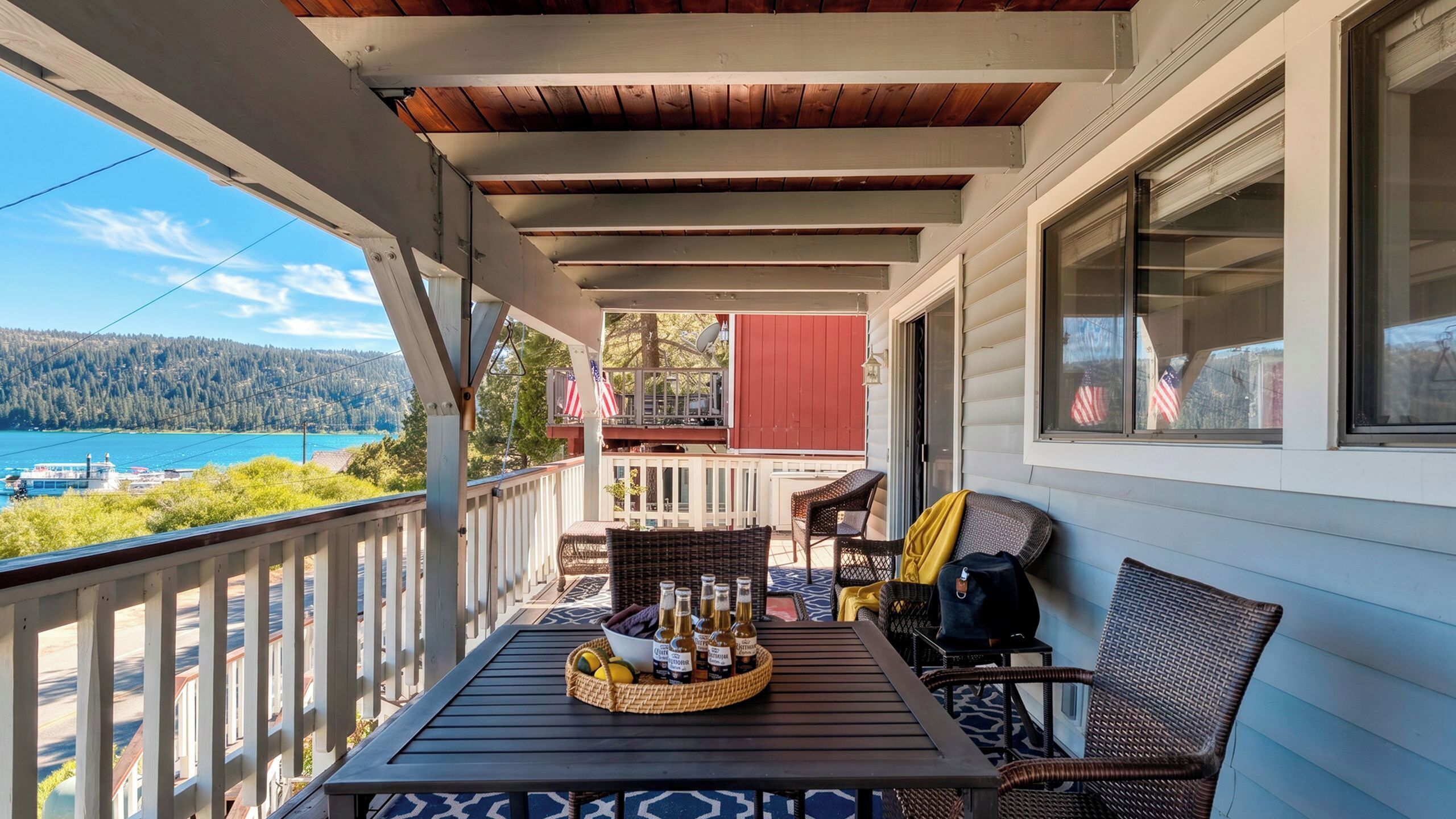 Un porche cubierto con una mesa lista para bebidas, sillas de mimbre y vistas a un lago y un bosque.