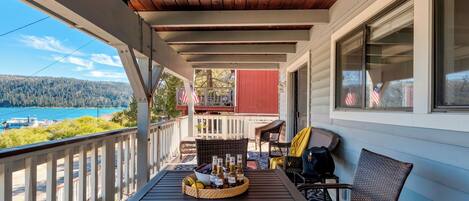 A covered porch with a table set for drinks, wicker chairs, and a view of a lake and forest.
