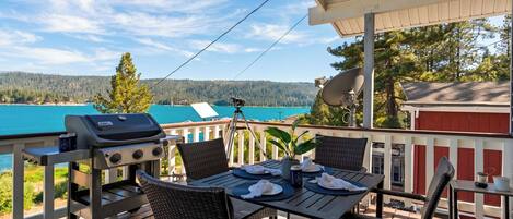 Una terraza con una parrilla, mesa de comedor, sillas y una vista panorámica de un lago y un bosque.