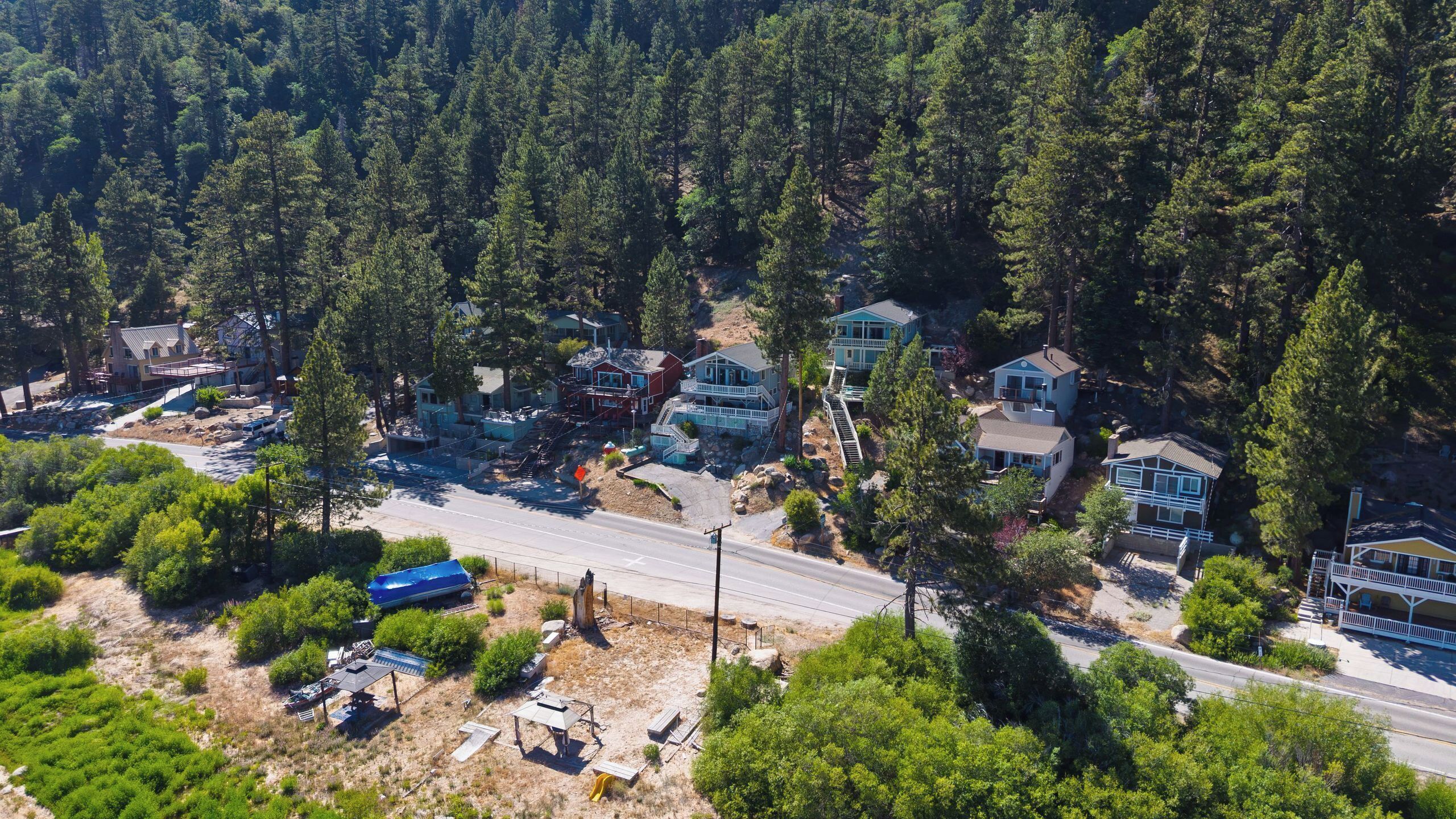 An aerial view of a residential area with houses surrounded by dense forest, a main road, and some undeveloped land.