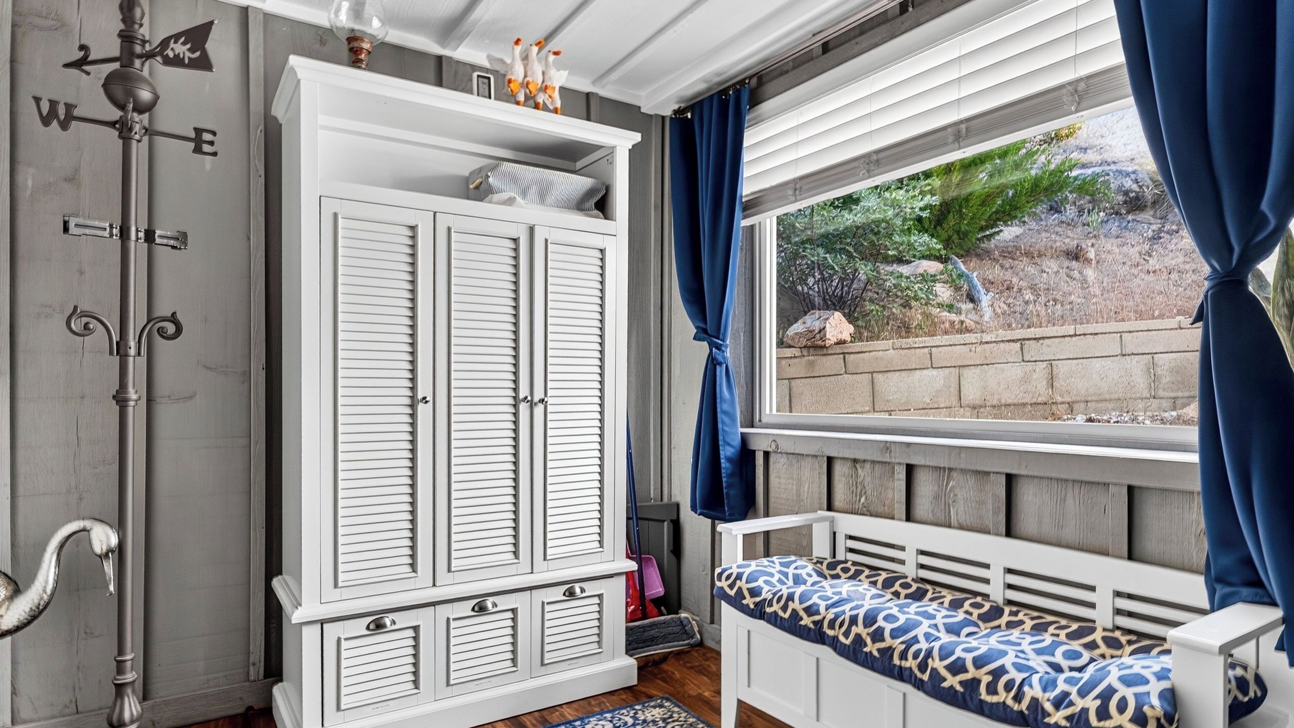 A room with a white wardrobe, a bench with a patterned cushion, a window with blue curtains, and a view of a garden.