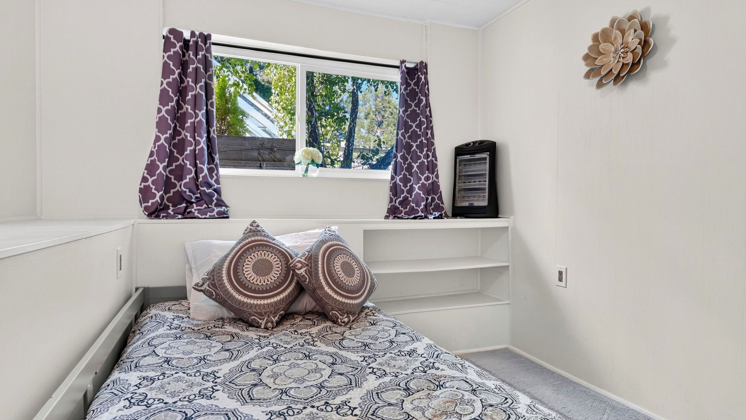 Un dormitorio con cama, ropa de cama estampada, una ventana con cortinas, un calentador y una decoración en la pared.