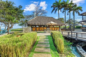 A thatched-roof building with a stone pathway leading to it, surrounded by lush greenery and palm trees.