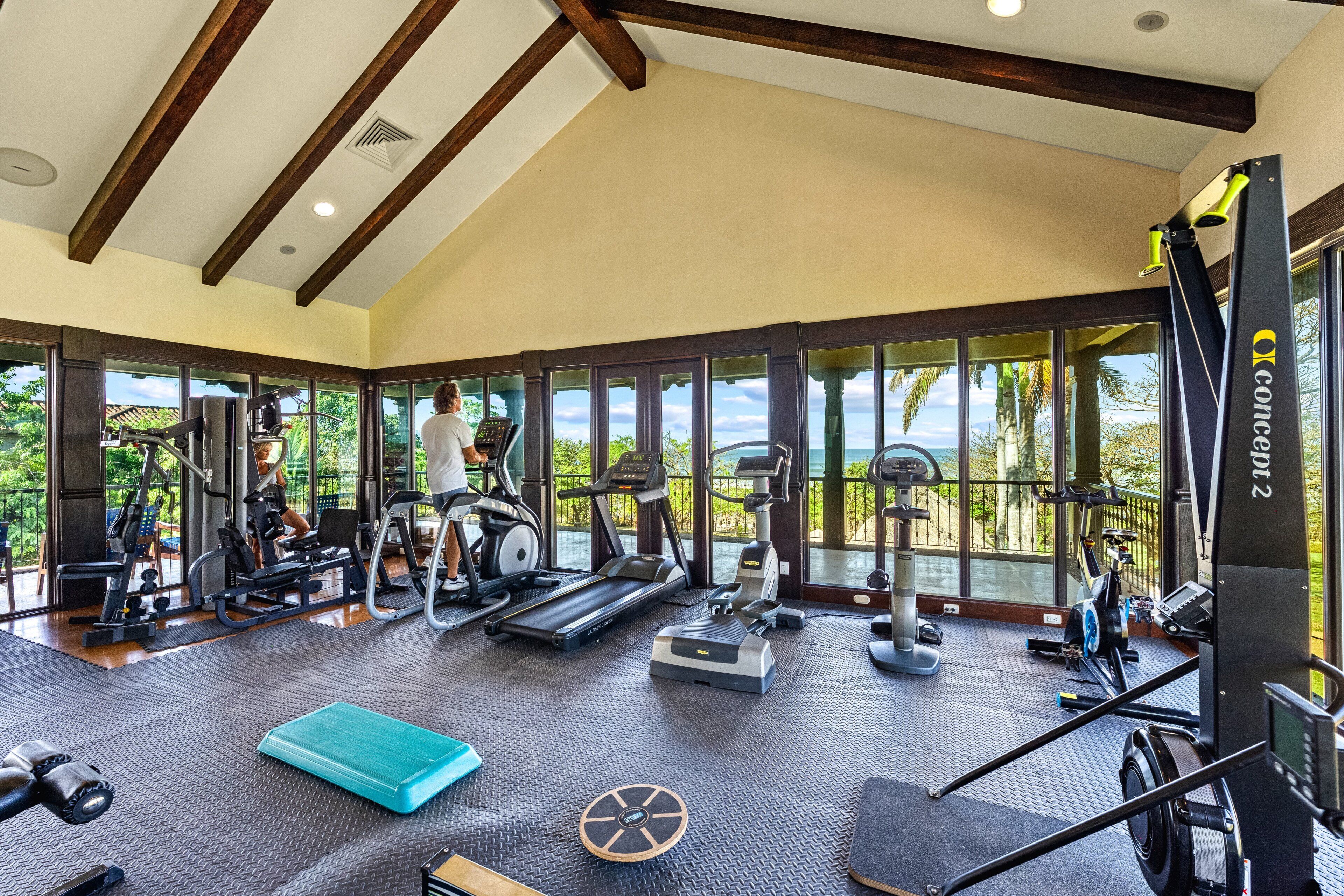 Ein Fitnessstudio mit Laufbändern, Crosstrainern und Kurzhanteln, großen Fenstern und einer hohen Decke mit Holzbalken.