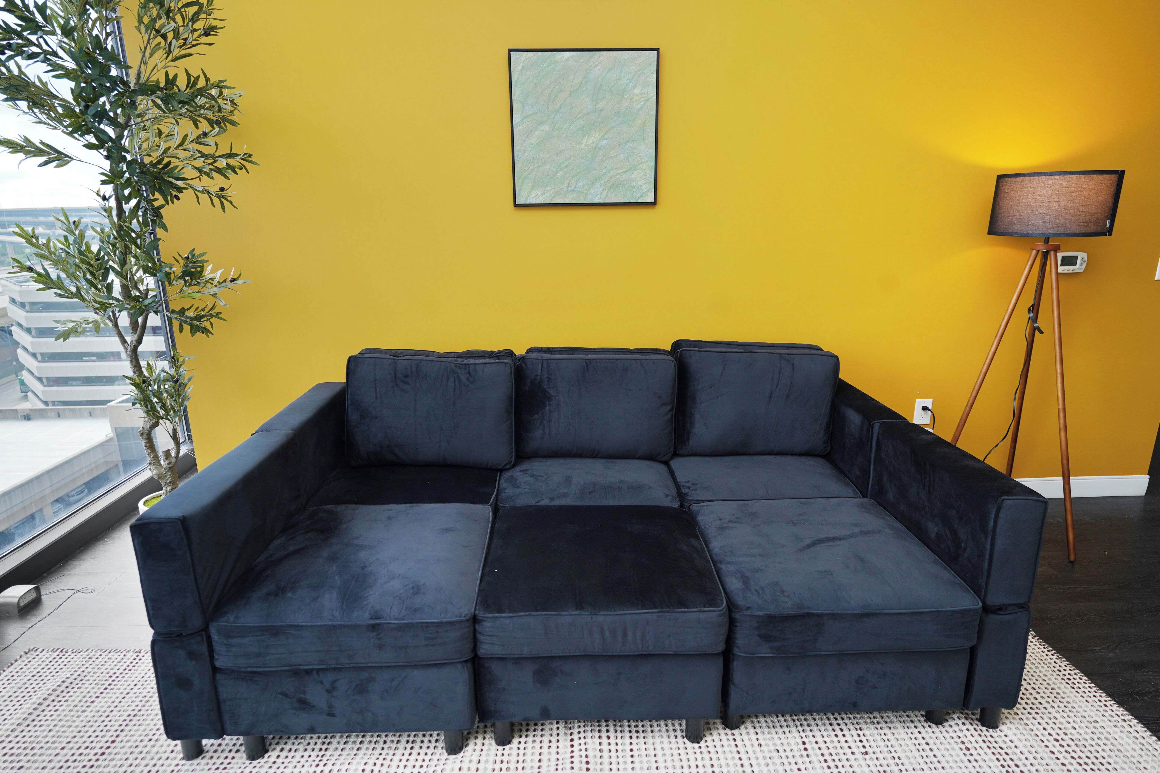 A black sectional sofa in a room with a yellow wall, a floor lamp, and a potted plant.