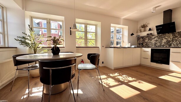 A modern kitchen with a round dining table, two chairs, a built-in oven, and a window overlooking a red building. - Heinason Collection - Old town & harbour (Thorshavn)