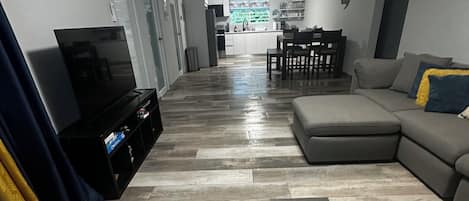 A living room with a flat-screen TV, a sofa, a dining table, and a kitchen in the background.