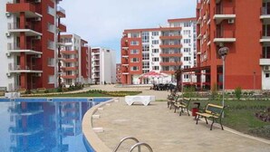 A swimming pool with a metal ladder, a paved walkway, and red and white apartment buildings in the background. - Grand Resort Marina View Fort Beach (Nessebar)