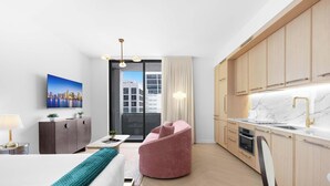 Ein modernes Hotelzimmer mit Bett, Schreibtisch, einem pinken Stuhl, einer Küche mit Holzschränken, einer Marmorarbeitsplatte und Blick auf die Stadt.