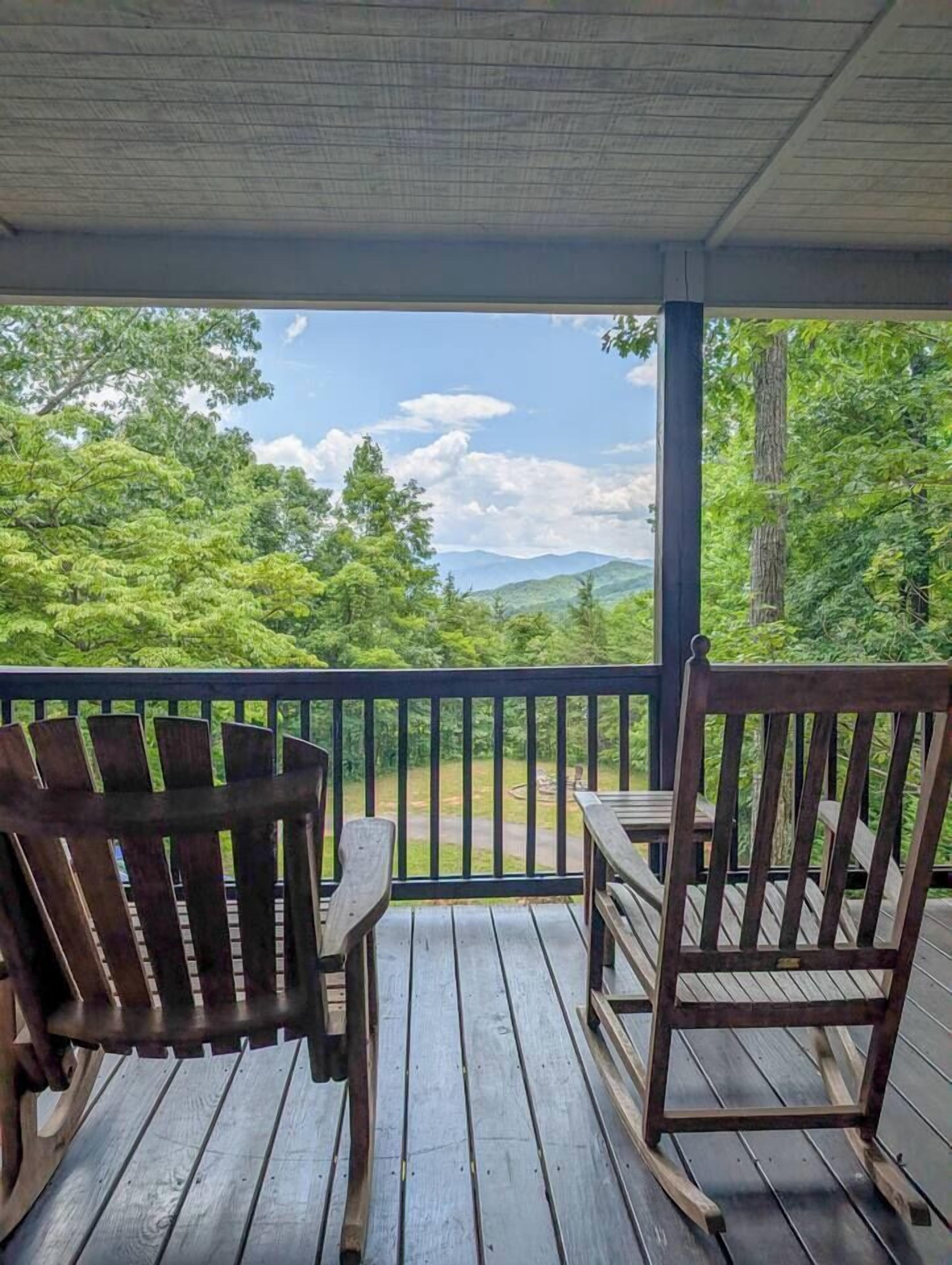 A wooden deck with two chairs and a table, overlooking a forested landscape with mountains in the distance.