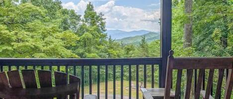 A wooden deck with two chairs and a table, overlooking a forested landscape with mountains in the distance.