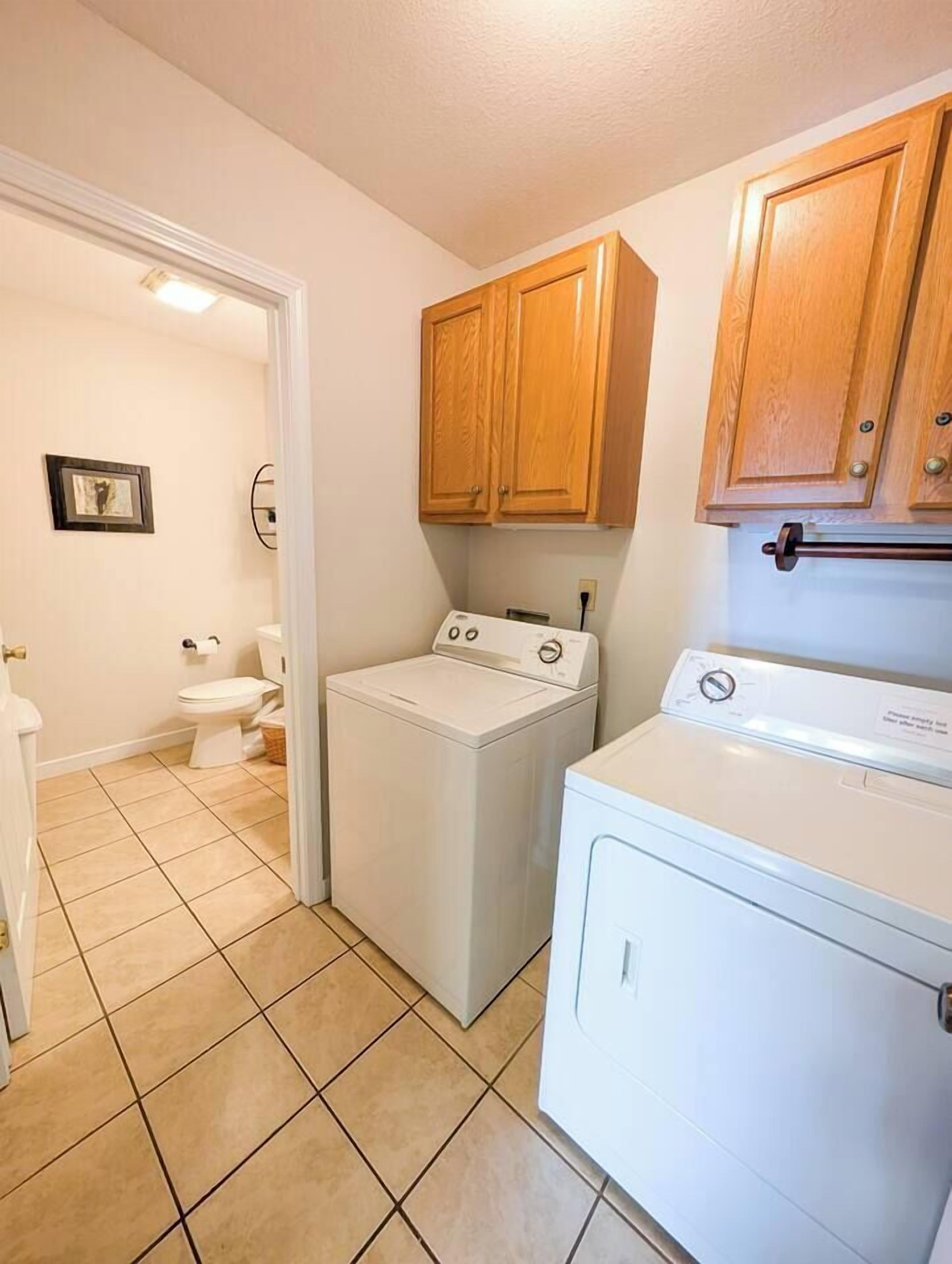 A laundry room with a washer and dryer, wooden cabinets, and a toilet in the background.